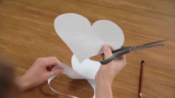 Una mujer joven dibuja un corazón en un papel blanco y luego lo corta con tijeras. Producción de regalos por amor — Vídeos de Stock
