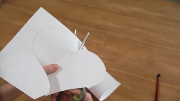 Una giovane donna disegna un cuore su una carta bianca e poi lo taglia con le forbici. Produzione di regali per amore — Video Stock