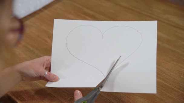 Uma jovem mulher desenha um coração em um papel branco e, em seguida, corta-o com tesoura. Produção de presentes por amor — Vídeo de Stock