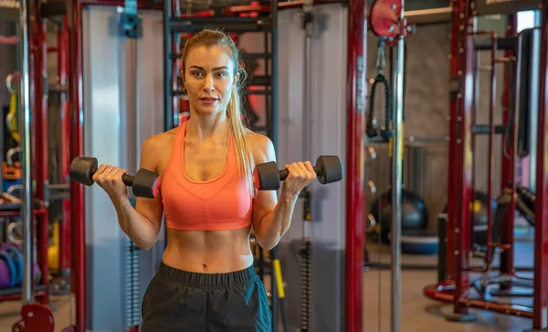 Mujer joven haciendo ejercicio con pesas para fortalecer los bíceps y los hombros en el gimnasio — Foto de Stock