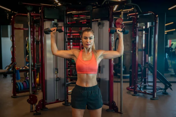 Mujer joven haciendo ejercicio con pesas para fortalecer los bíceps y los hombros en el gimnasio — Foto de Stock