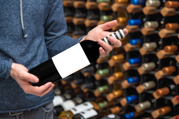 man in a wine shop holding a bottle with a blank label in his hand. A rack with wine in the background.