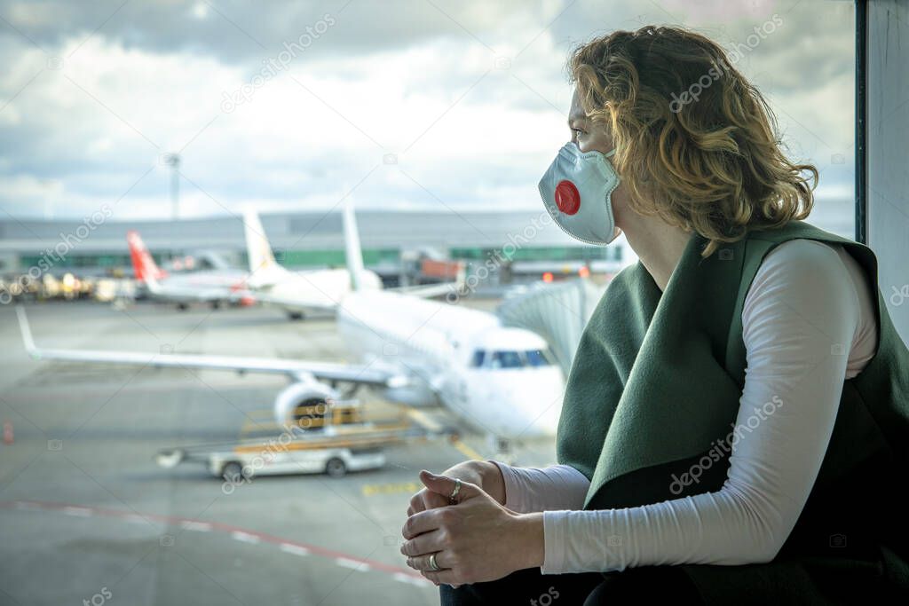 Woman with a mask on her mouth protects against the virus. She looks sad through the window at the airport on planes. Aerial connections canceled due to a coronavirus