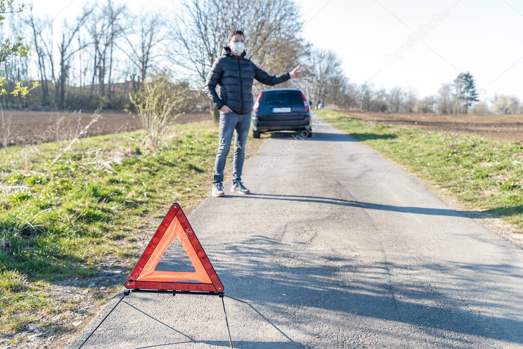 red warning triangle on the road in front of a broken car. The young man is looking for help stopping vehicles moving around