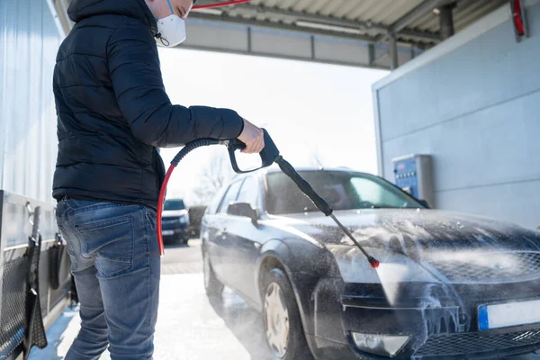 Washing car by water pressure at self-service station. A man with a respirator on his face protects himself from a viral epidemic — Stock Photo, Image