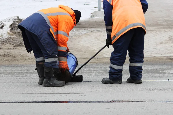Dos trabajadores de la carretera en overoles se vierten con alquitrán caliente en el borde de la tira de la carretera al reparar la carretera — Foto de Stock
