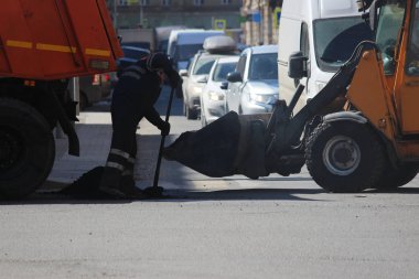 Çöp kamyonu kazıcının kovasına asfalt kırıntıları döktü. Makinelerin hareketi sırasında aşırı yol onarımı olması ihtimaline karşı. Kürek kullanan bir işçi kazıcının kovasından bir çukuru doldurur..