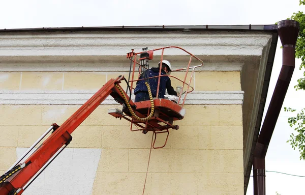 Um pintor jovem do trabalhador pinta a linha branca da parede do edifício da estação ferroviária da cidade na altura com a ajuda de um elevador — Fotografia de Stock