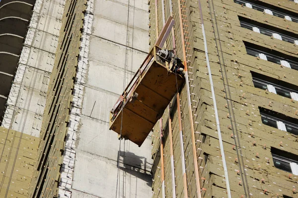 Worker in yellow suspended cradle mounts environmental boards for insulation on a newly built high-rise building — Stock Photo, Image