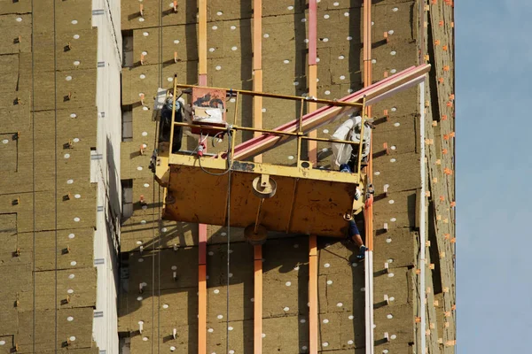 Arbeiter in gelber Hängewiege montiert Umweltplatten zur Isolierung an einem neu errichteten Hochhaus — Stockfoto