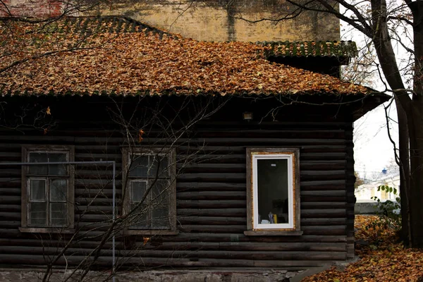 Ein altes Holzhaus, dessen Dach mit Herbstblättern bedeckt ist. eine Kombination aus Antike und einem neuen weißen Kunststofffenster — Stockfoto