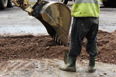 Traktör bir kova ile toprak parçası düzeltme eki için yolun asfaltlama önce kaldırır. Foreman inşaatçılar yan yana duruyor ve ekskavatör çalışmaları yönetiyor.