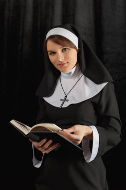 young nun in a robe holding  bible and read against the dark wall. Close-up. Woman looking at the camera clipart
