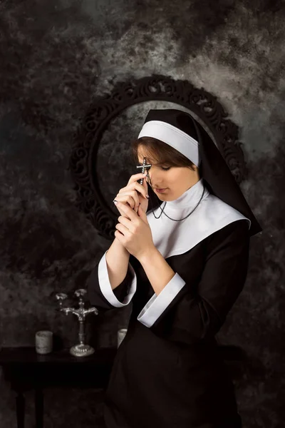 Monja joven con túnicas sosteniendo la cruz y rezando sobre el fondo oscuro de la pared — Foto de Stock