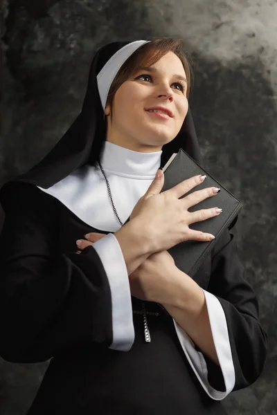 Monja joven con una túnica que sostiene la Biblia y se cruza contra la pared oscura. Primer plano. Libro de abrazos de mujer — Foto de Stock