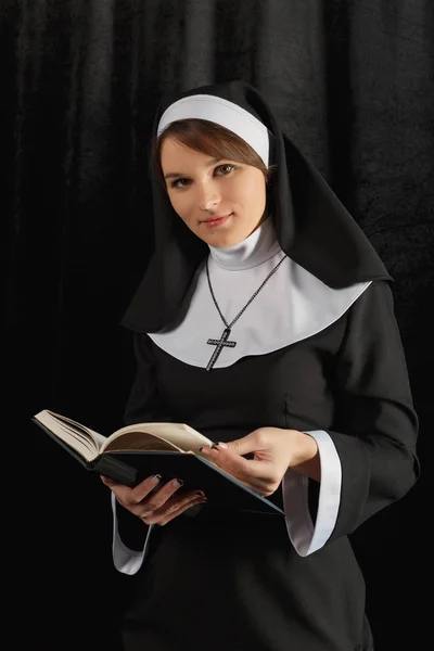 Monja joven con una túnica que sostiene la Biblia y lee contra la pared oscura. Primer plano. Mujer mirando a la cámara — Foto de Stock