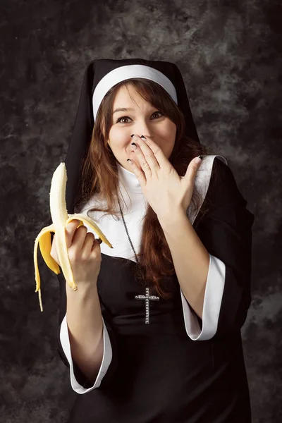 근접 한 바나나를 들고 섹시 한 수녀의 어두운 배경에 부끄러움에 그의 얼굴을 커버 하 고. 여자는 바나나를 먹는다 — 스톡 사진