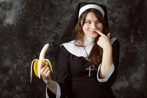 어두운 배경에 그의 코 바나나와 카트리지 엄지손가락을 들고 섹시 한 수녀의 클로즈업. 여자는 바나나를 먹는다 — 스톡 사진