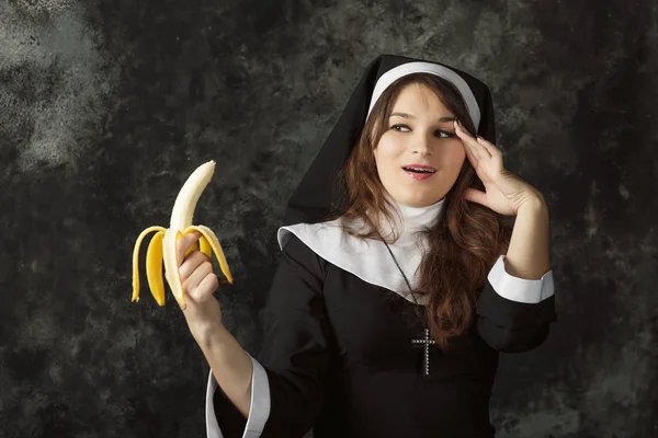 Close-up de uma freira sexy segurando uma banana e segurando a cabeça na vergonha em um fundo escuro. Mulher come banana — Fotografia de Stock