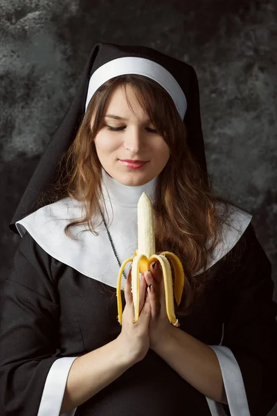 Gros plan d'une nonne sexy tenant la main banane EYAD sur un fond sombre. Une femme mange une banane. Une religieuse prie avec de la banane — Photo