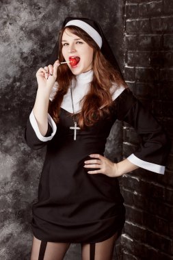 young sexy nun in stockings licking a red candy on a dark background.Attractive woman enjoys eating bonbon clipart