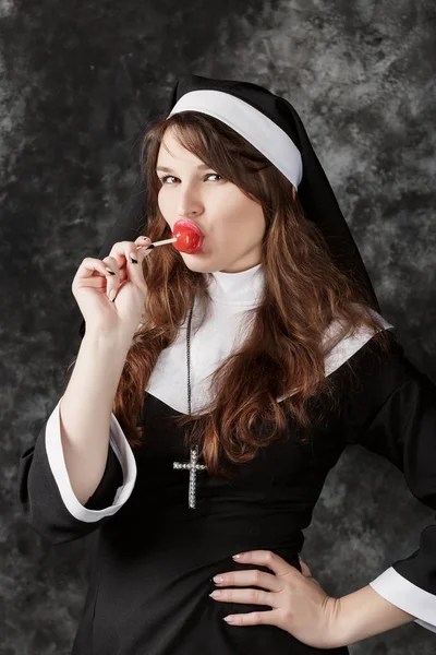 Young sexy nun in stockings licking a red candy on a dark background.Attractive woman enjoys eating bonbon — Stock Photo, Image