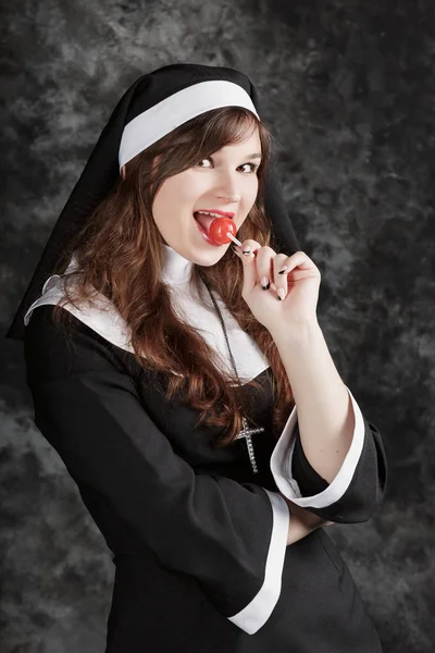 Monja sexy joven en medias lamiendo un caramelo rojo en un fondo oscuro. Mujer atractiva disfruta comiendo bonbon — Foto de Stock