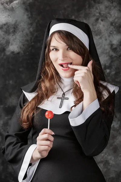 Joven sexy medias monja sosteniendo un rojo bonbon en un oscuro background.attractive chica morena con caramelo lamiendo dedo . — Foto de Stock