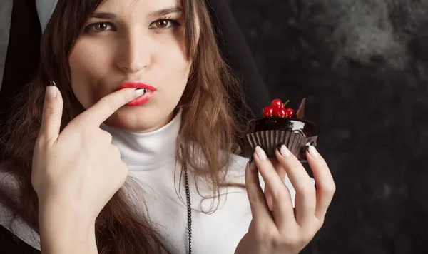 Closeup freira atraente segurando um cupcake e lambendo o dedo em um fundo escuro — Fotografia de Stock