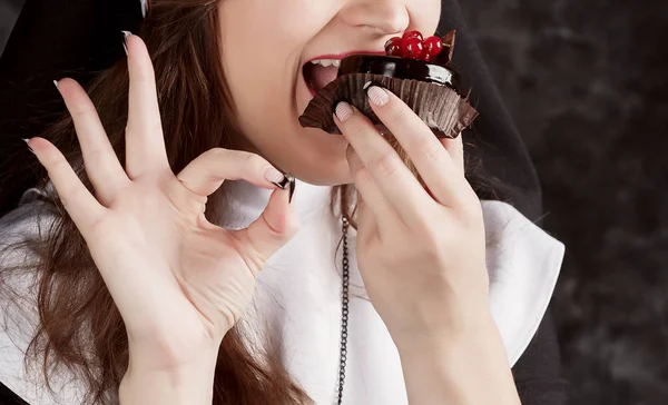 Uma freira em uma mão segurando um pedaço de bolo de chocolate com groselhas e morde-o em um fundo escuro . — Fotografia de Stock