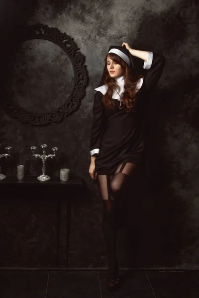 Atractiva monja sexy en medias y zapatos se encuentra cerca de una pared en una habitación oscura —  Fotos de Stock