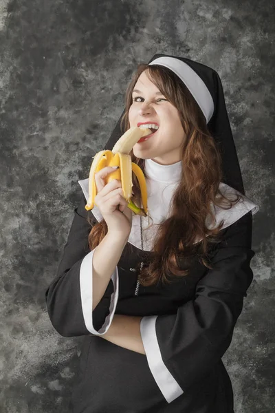 Freira segurando uma banana e morde-o em um fundo escuro — Fotografia de Stock