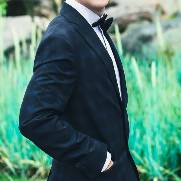 Closeup groom in a blue suit, white shirt and tie butterfly holds hands in pockets on a background of a green bush — Stock Photo, Image