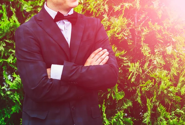 Closeup groom in a blue suit, white shirt and tie butterfly arms crossed against a background of a green bush. — Stock Photo, Image