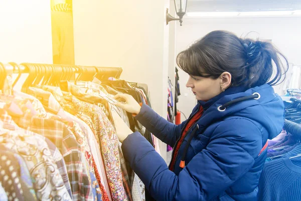 Młoda brunetka Kobieta w Błękitnej Kurtki wybiera ubrania w sklepie. Kobieta kupuje ubrania — Zdjęcie stockowe