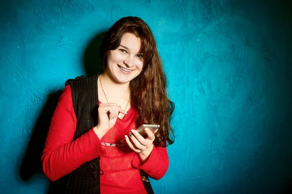 Brunette woman in a red blouse and black jacket standing against the backdrop of a blue wall and use a smartphone — Stock Photo, Image