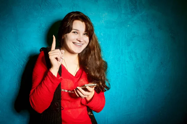 Brunette woman in a red blouse and black jacket standing against the backdrop of a blue wall and use a smartphone — Stock Photo, Image
