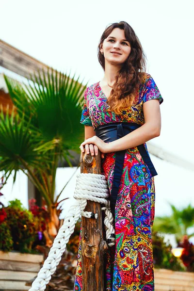 Bela morena em um vestido longo colorido em pé na praia perto do bar no fundo de palmeiras . — Fotografia de Stock