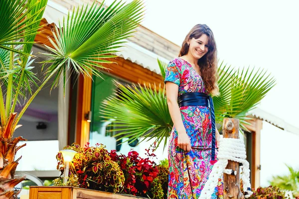 Hermosa morena en un vestido largo de colores de pie en la playa cerca del bar en el fondo de las palmeras . — Foto de Stock