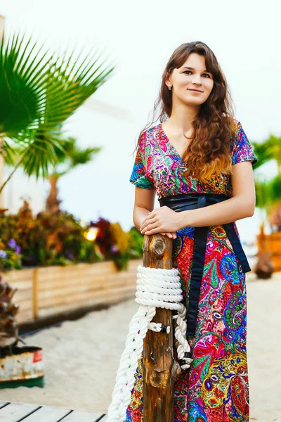 Hermosa morena en un vestido largo de colores de pie en la playa cerca del bar en el fondo de las palmeras . — Foto de Stock