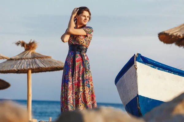 Atractiva morena en vestido largo y colorido de pie solo en la playa cerca del barco al atardecer — Foto de Stock
