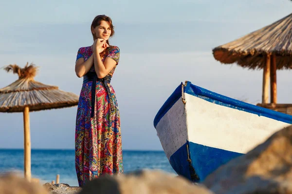 Atractiva morena en vestido largo y colorido de pie solo en la playa cerca del barco al atardecer — Foto de Stock