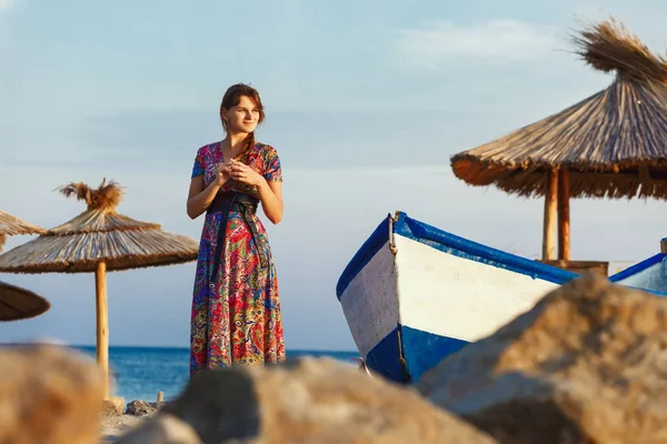 Atractiva morena en vestido largo y colorido de pie solo en la playa cerca del barco al atardecer — Foto de Stock