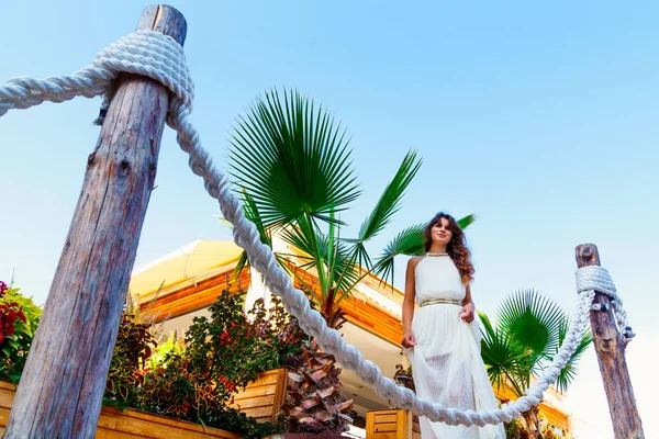 Atractiva mujer morena en un vestido blanco de pie sobre el fondo del edificio y palmeras cerca de la cuerda de la valla — Foto de Stock