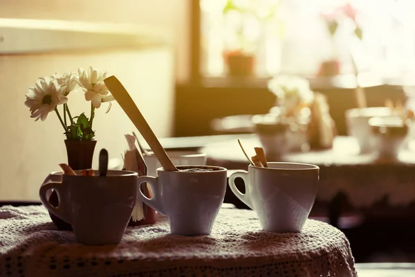 Stilleven. Witte porseleinen kopjes en een vaas met bloemen staan op een tabel met een wit tafellaken in een bruin café-zaal — Stockfoto