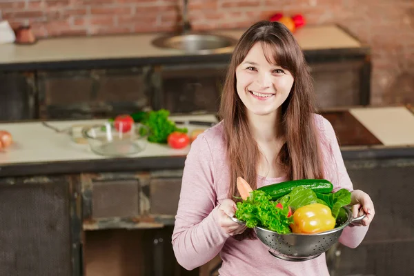 La morena en la cocina sostiene un cuenco con verduras frescas y mira a la cámara y sonríe. Alimento saludable . — Foto de Stock