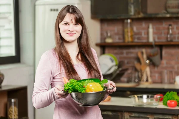 La morena en la cocina sostiene un cuenco con verduras frescas y mira a la cámara y sonríe. Alimento saludable . — Foto de Stock