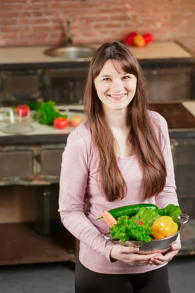 Bruneta žena v kuchyni drží misku s čerstvou zeleninou a dívá na kameru a úsměvy. Zdravé jídlo. — Stock fotografie