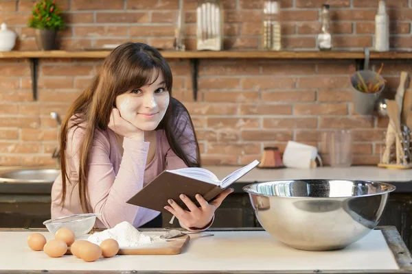 En kvinna förbereder mat i köket och läser matlagning recept i boken. — Stockfoto