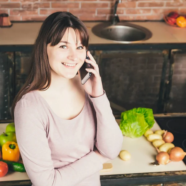 Sonriente joven mensajería de texto frente a las verduras en la cocina en casa — Foto de Stock